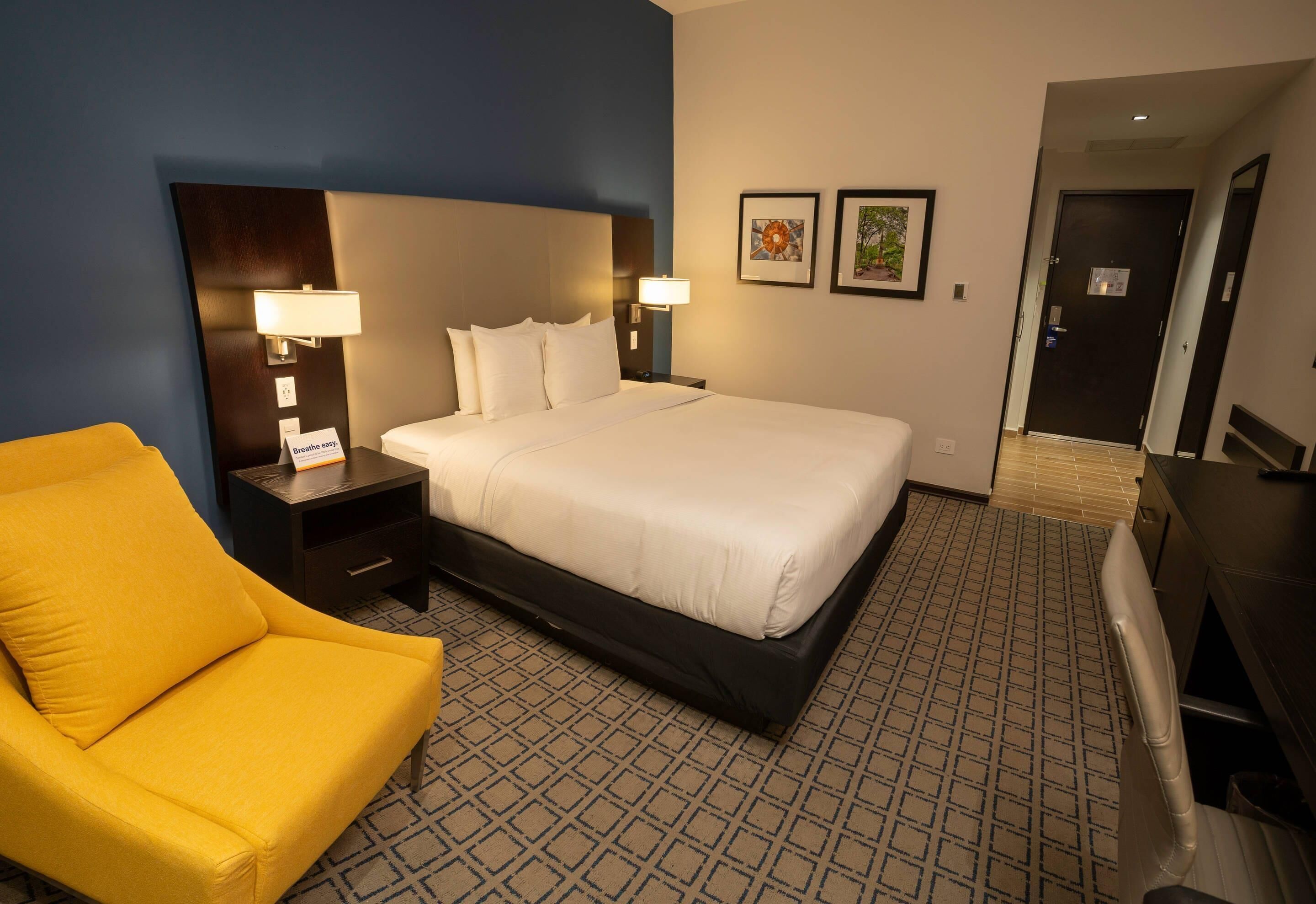 A hotel room with a large bed, a yellow armchair, a desk, and two wall-mounted lamps.