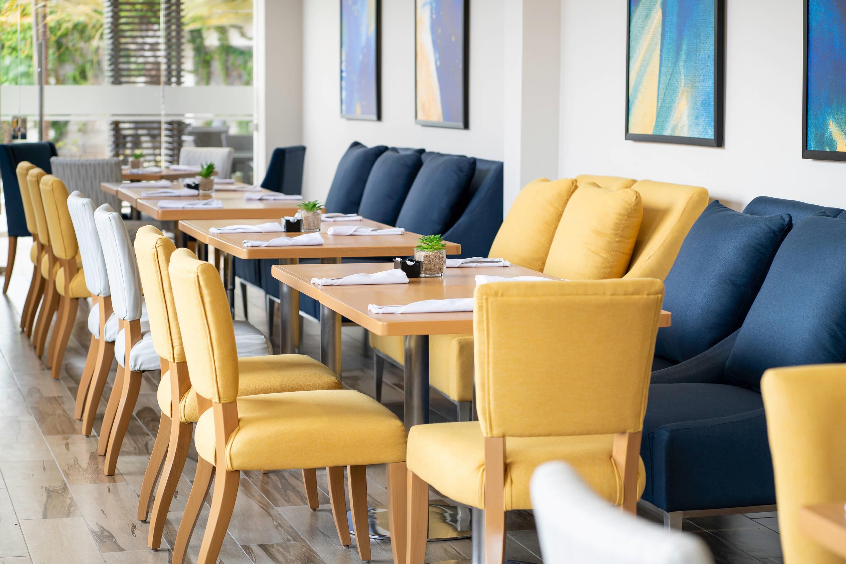 A modern dining area with long tables and yellow chairs, blue sofas, and artwork on the walls.