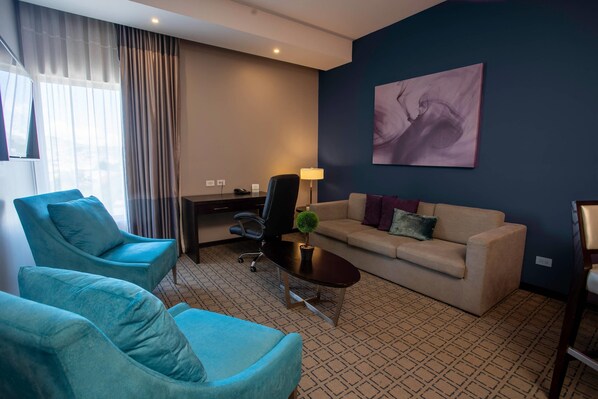 A hotel room with a blue armchair, a beige sofa, a small table with a lamp, and a painting on the wall.