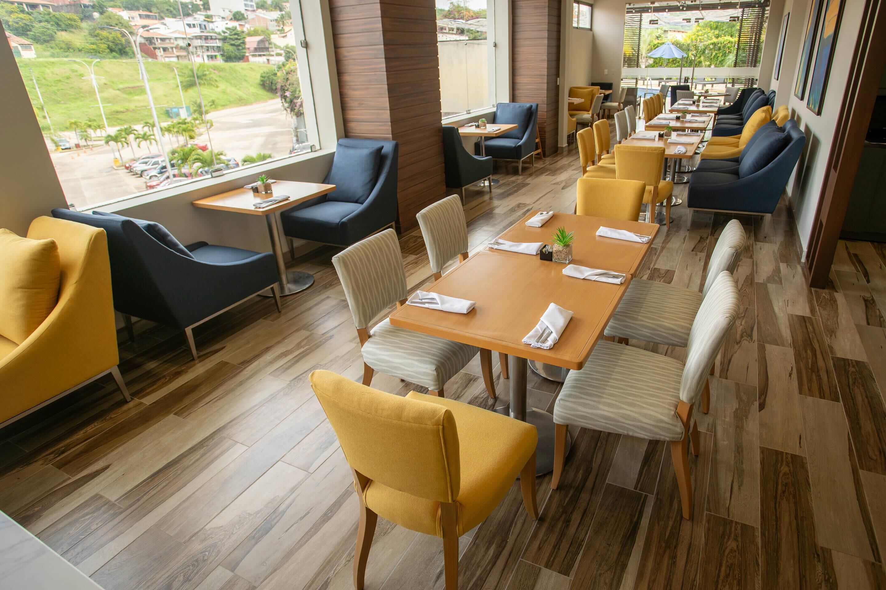 Ein modernes Restaurant mit Holzboden, gelb-blauen Sitzmöbeln und großen Fenstern mit Blick auf die Straße.