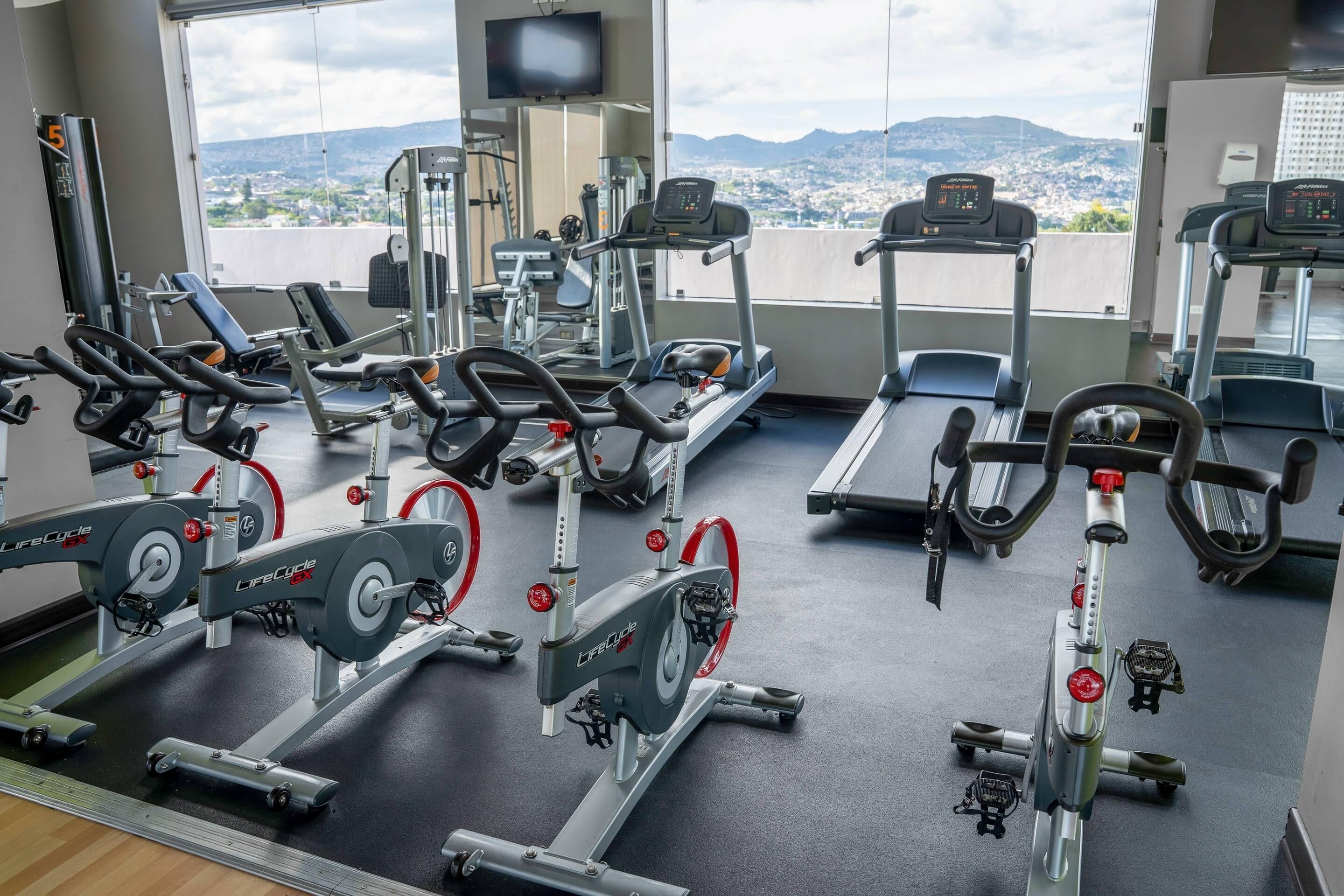 Ein Fitnessstudio mit unterschiedlichen Trainingsgeräten, einem großen Fenster mit Blick auf die Stadt und einem an der Wand montierten Fernseher.