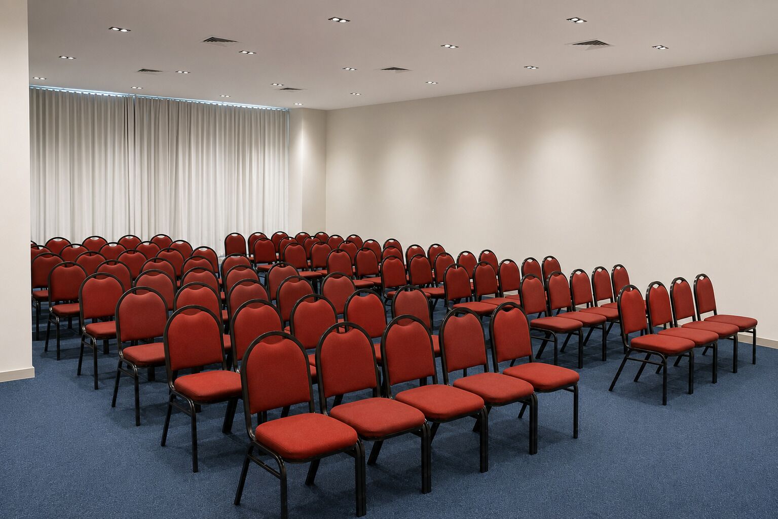 Un salón de conferencias con hileras de sillas rojas y mesas, una alfombra azul y un fondo de cortina blanca.