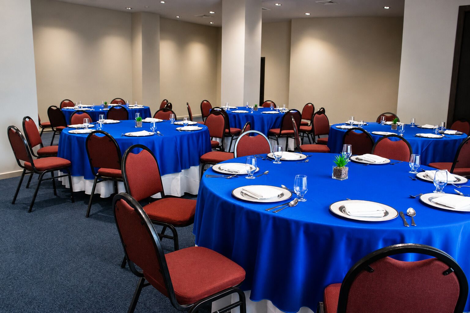 Un salón de conferencias con varias mesas redondas, cada una con platos blancos, cubiertos y vasos transparentes.
