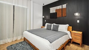 Une chambre moderne avec un lit en bois, une literie blanche, une couverture rayée grise et blanche, deux tables de chevet avec des lampes et un mur noir à texture.