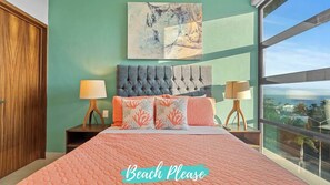 Une chambre avec un grand lit, deux tables de chevet avec des lampes, une vue sur l’océan et un tableau au mur.