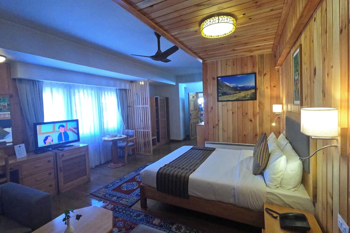 A wooden-themed hotel room with a bed, bedside lamp, TV, desk, and a view of a mountainous landscape.