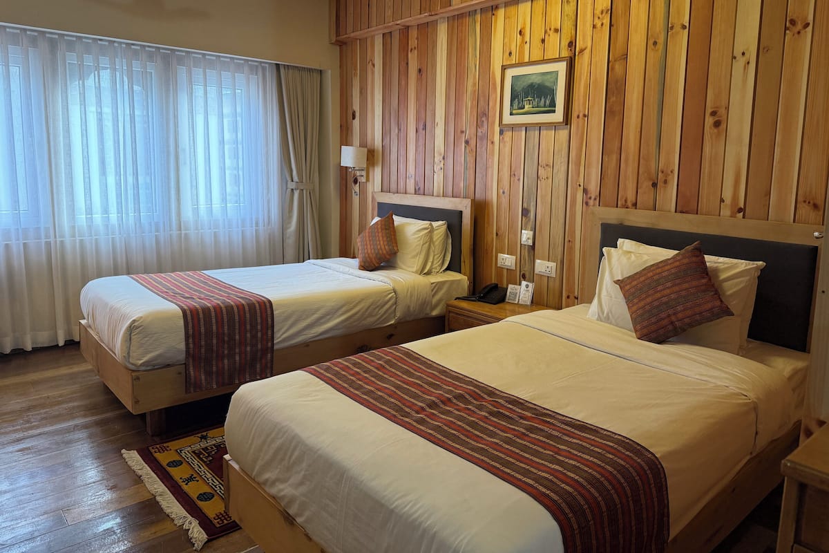 A room with two beds, wooden paneling, a nightstand with a lamp, and a window with sheer curtains.