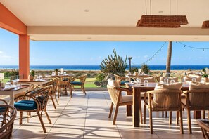 An outdoor dining area with tables and chairs, overlooking a beach and ocean view.