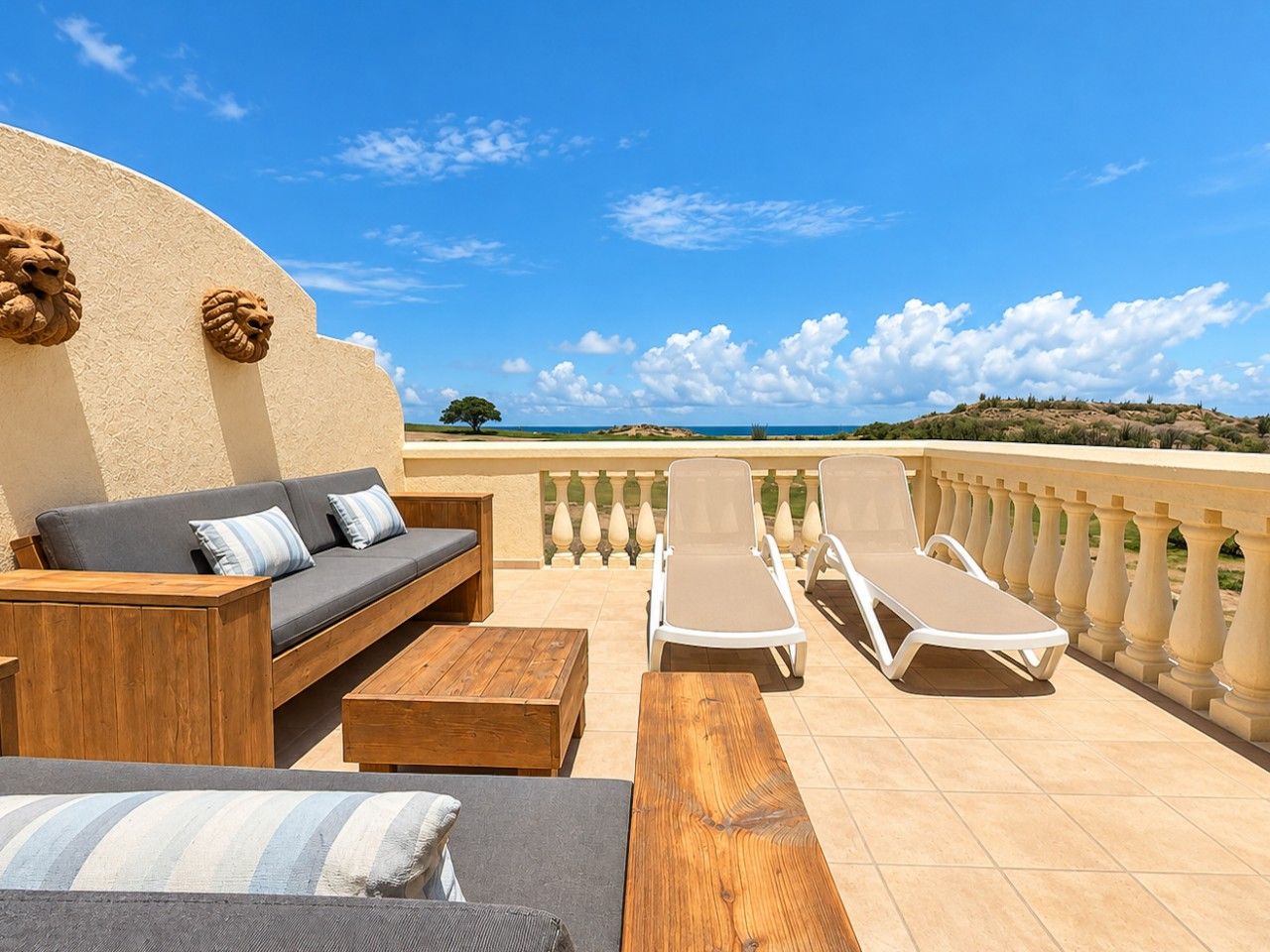 A sunlit outdoor seating area with a sofa, lounge chairs, and a wooden table, overlooking a scenic landscape.