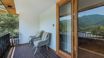 A balcony with wooden flooring, two wicker chairs, and a view of a mountainous landscape through a glass door.