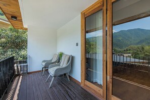 Un balcon doté d’un plancher en bois, de deux fauteuils en osier et offrant une vue sur un paysage montagneux, visible à travers une porte vitrée.