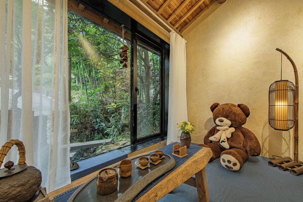 A room with a large window, a wooden bench, a teddy bear, and a traditional lamp.