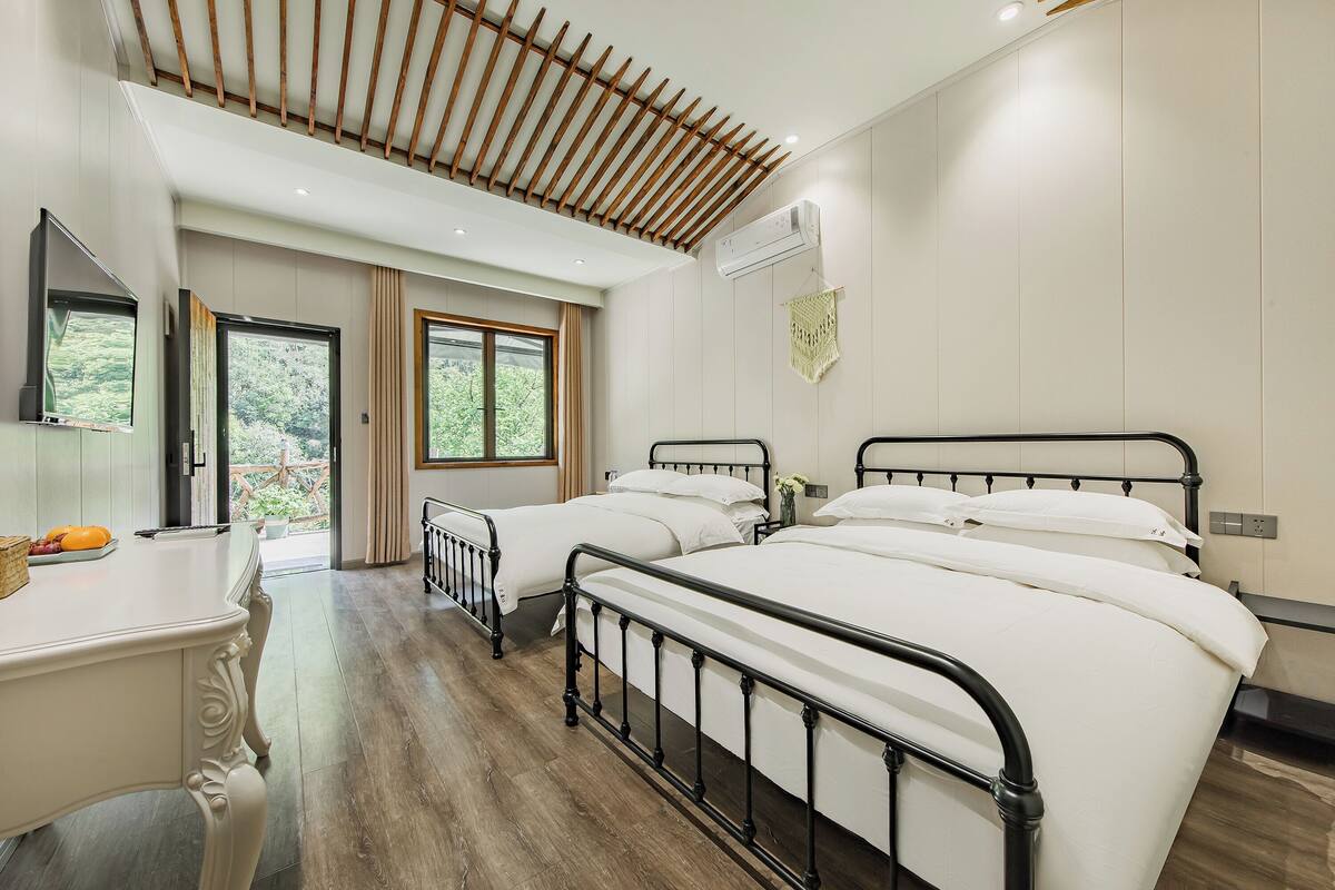A bedroom with two beds, a desk, and a window with a view of greenery.