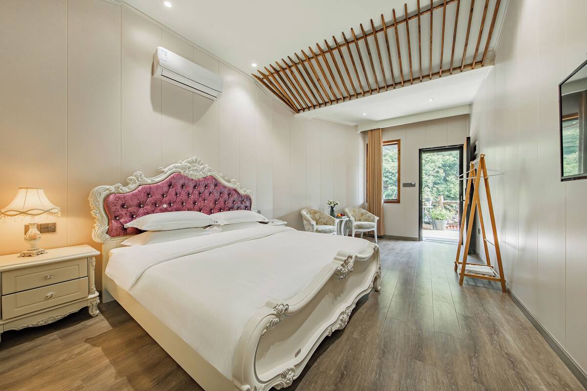 A modern bedroom with a large bed, a nightstand, a dresser, a wooden ladder, and a view of the outdoors.