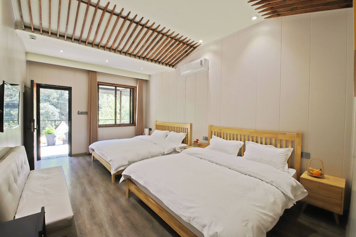 A hotel room with two beds, a wooden ceiling, an air conditioning unit, a small table with a basket, and a view of greenery outside.