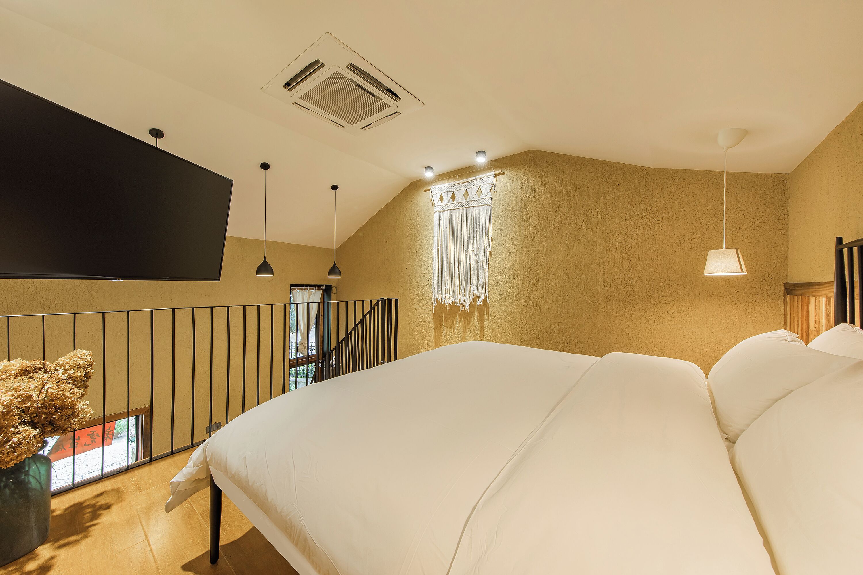 A bedroom with a large bed, a flat-screen TV mounted on the wall, a window with a view, and a staircase leading to an upper level.
