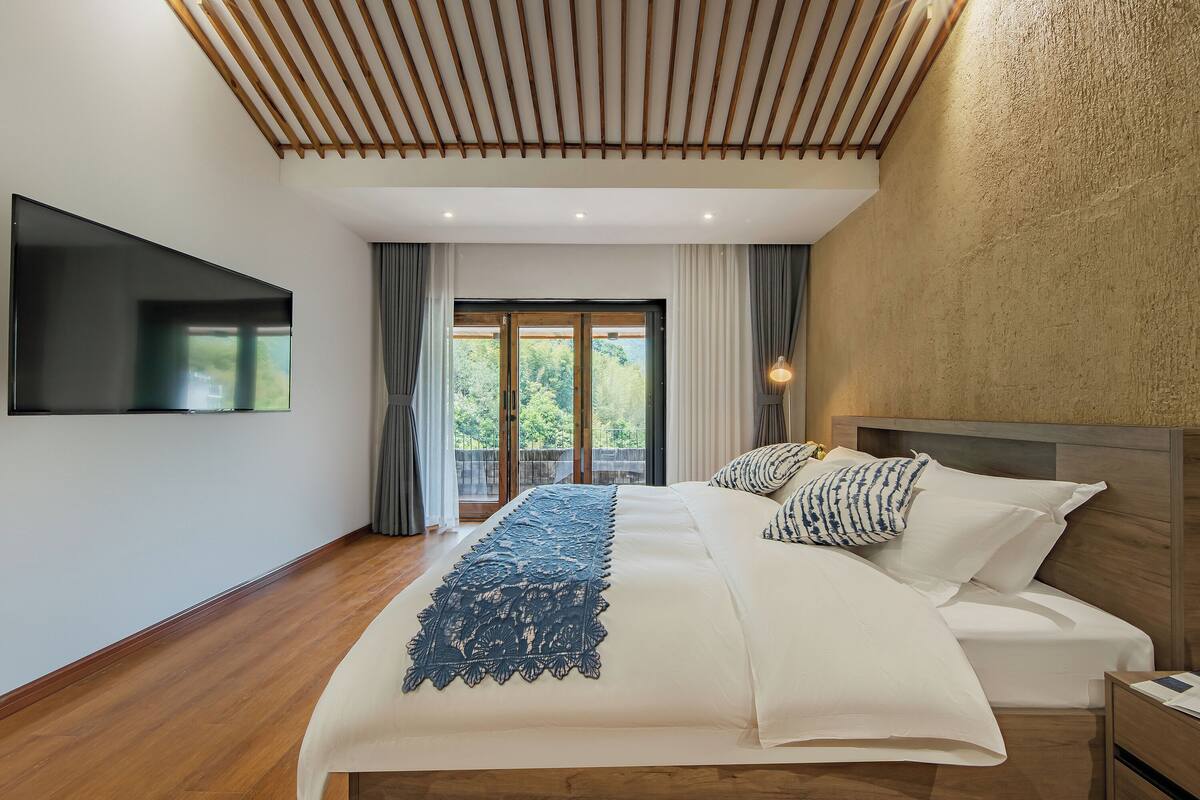 A modern bedroom with a large bed, a TV mounted on the wall, and a view of the outdoors through a sliding door.