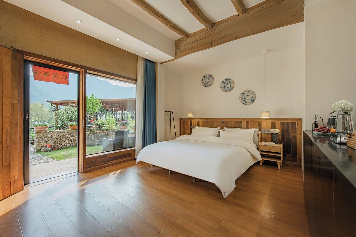 A spacious bedroom with a large bed, wooden beams, a view through a sliding door, and a small bedside table with a lamp.
