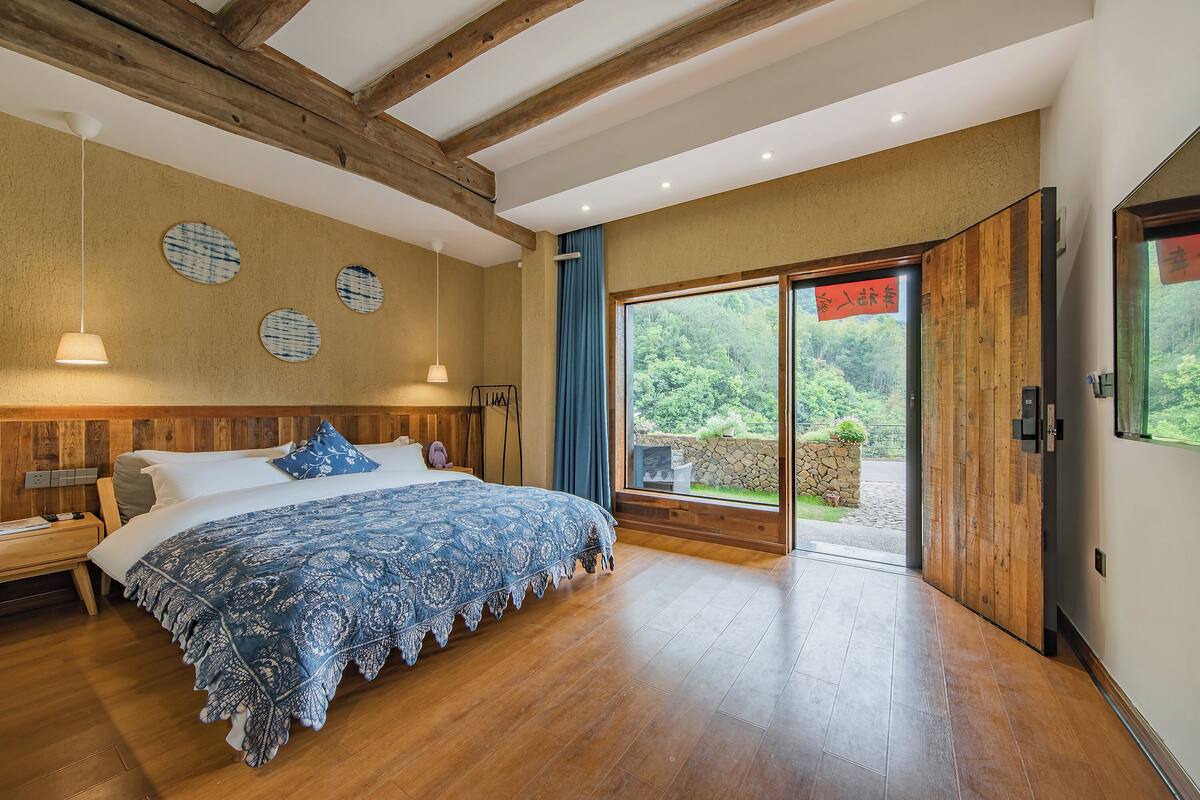 A bedroom with a wooden bed, a nightstand, a large window with a view of greenery, and a wooden ceiling.