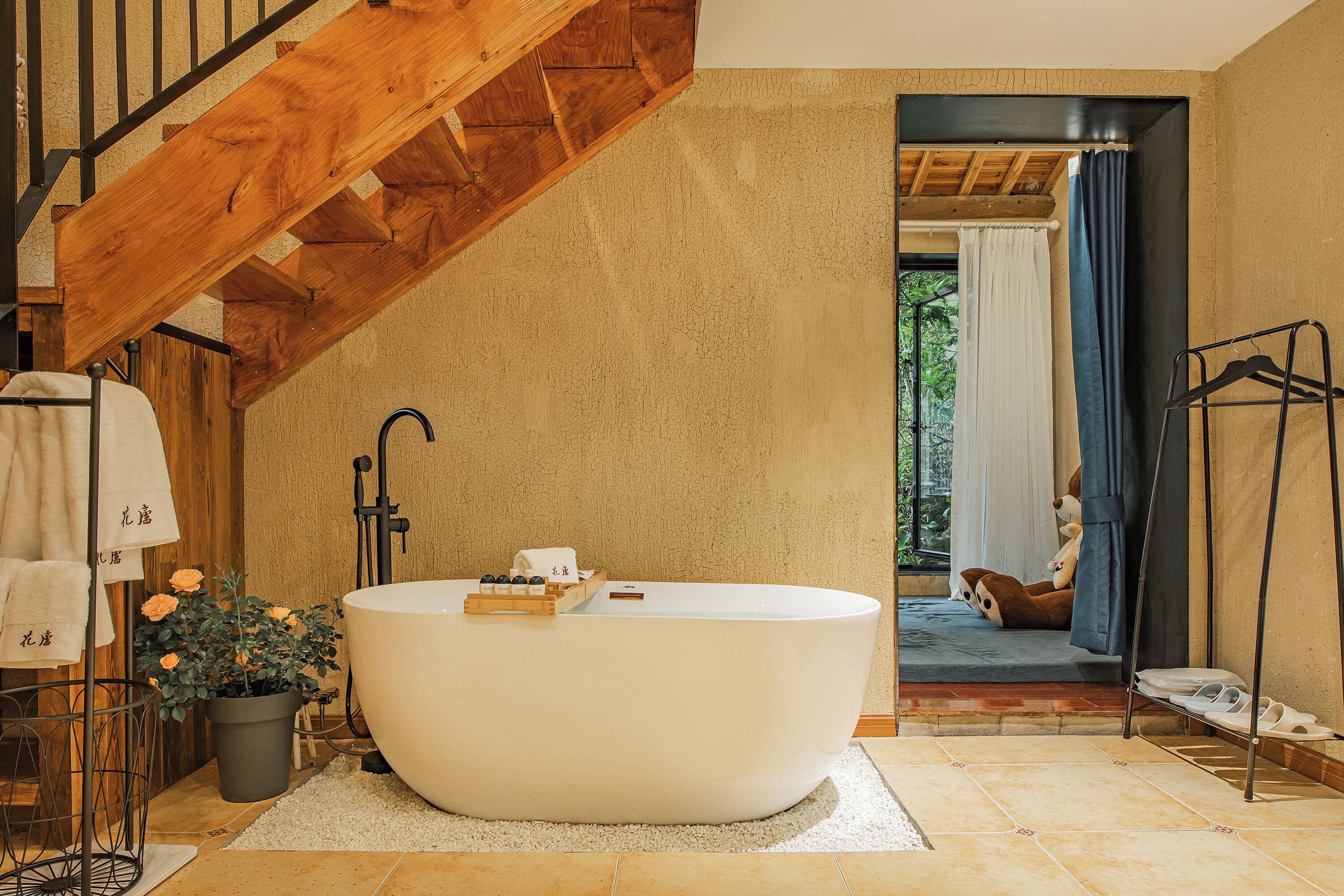 A modern bathroom with a freestanding bathtub, a wooden staircase, and a view of the outdoors through a large window.