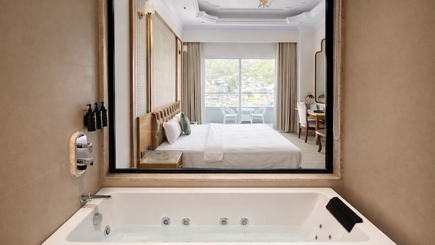 Ein Hotelzimmer mit großem Fenster, einer Badewanne, einem Bett mit weißer Bettwäsche und einem Schreibtisch mit Stuhl.
