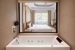 Une chambre d’hôtel avec une grande fenêtre, une baignoire, un lit avec du linge de lit blanc et un bureau avec une chaise.
