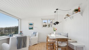 Un interior luminoso y blanco con suelo de madera, una mesa de comedor con sillas, un banco y una ventana con vistas a un bosque.
