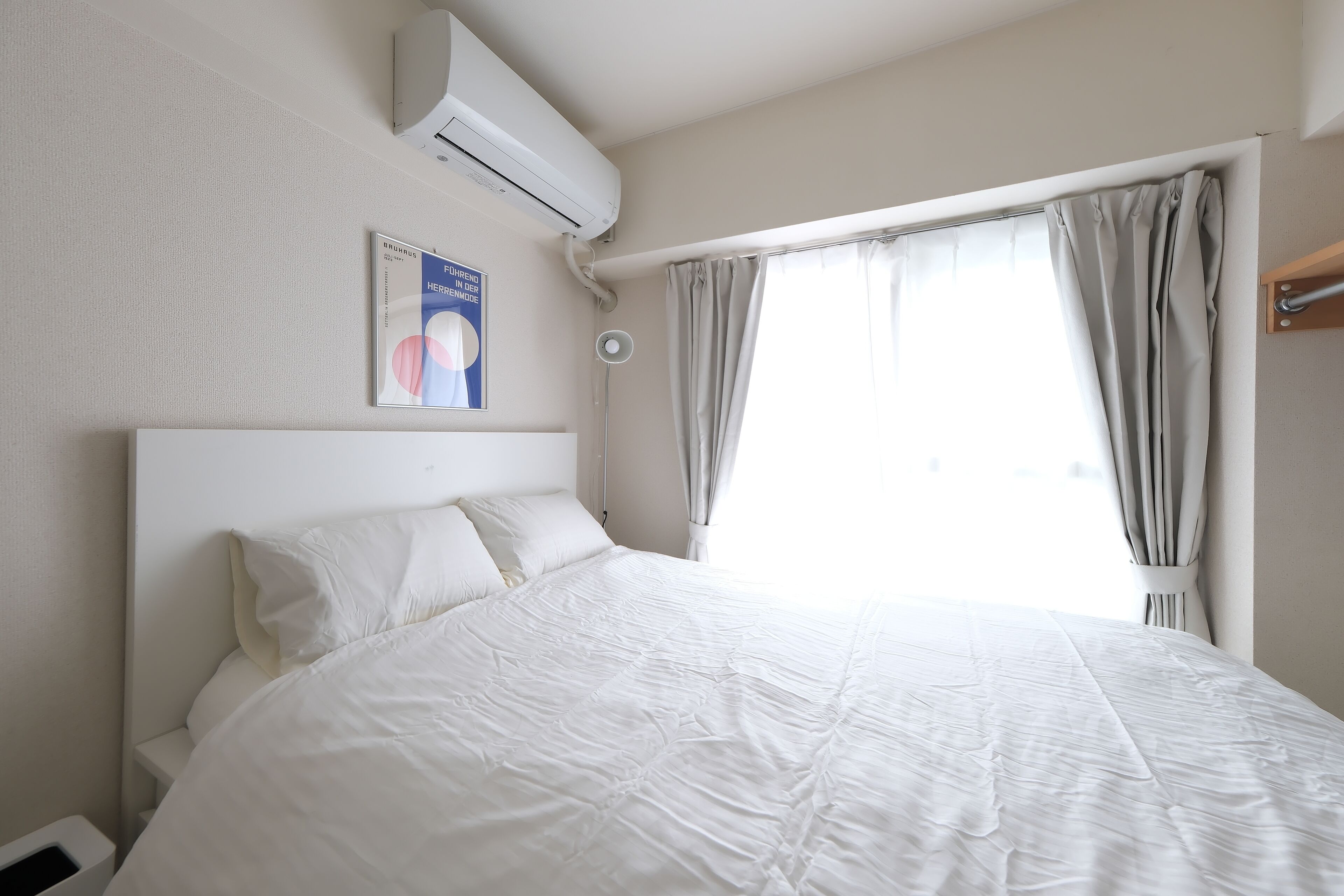 A hotel room with a large bed, an air conditioning unit, a bedside lamp, and a window with curtains.