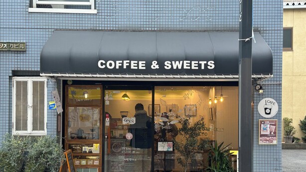 A coffee and sweets shop with a black awning, a wooden bench outside, and potted plants.