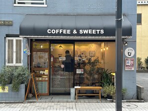 Una tienda de café y dulces con un toldo negro, una banca de madera afuera y plantas en macetas.