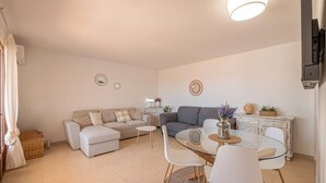 A modern living room with a sofa set, a glass dining table with chairs, a small side table with a vase, and a white cabinet with decorative items.