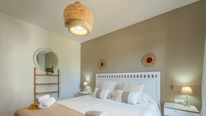 A neatly made bed with white and beige bedding, a bedside table with a lamp, and a hanging light fixture.