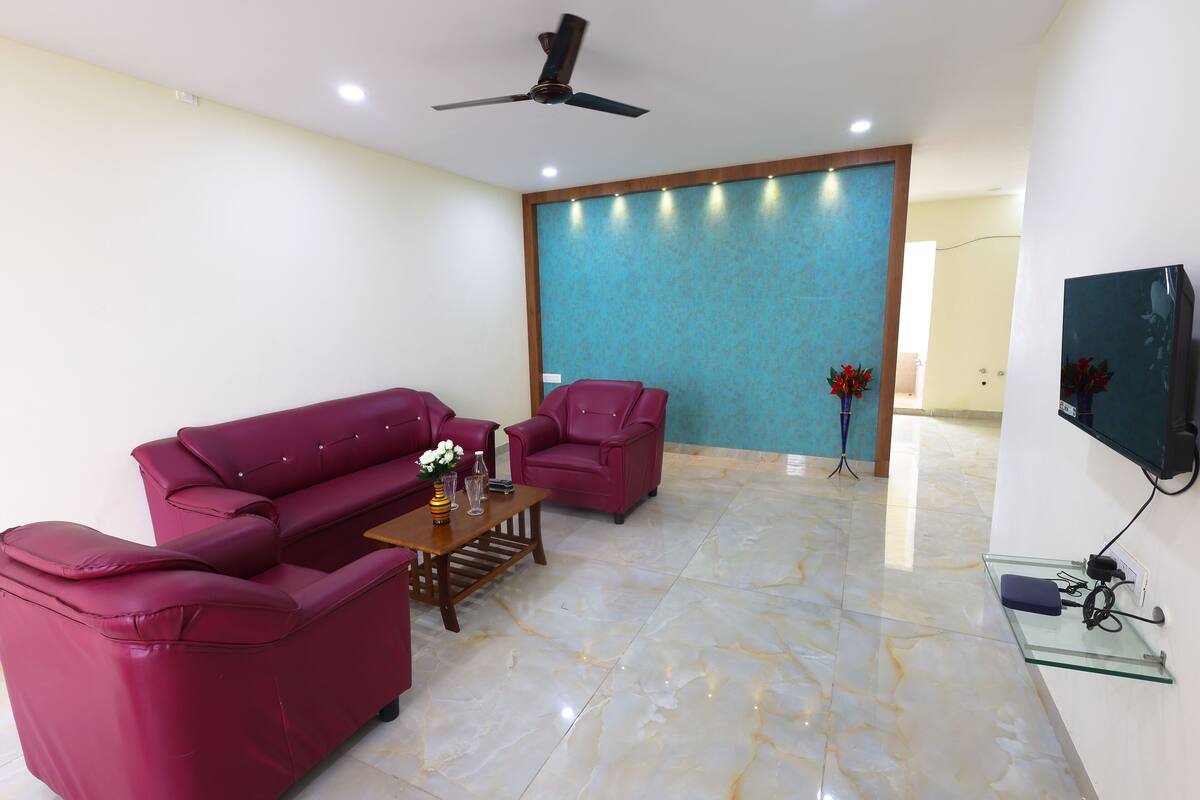 Uma sala de estar com sofá, poltrona e mesa de centro. Há um ventilador de teto, uma TV fixada na parede e uma parede azul decorativa.