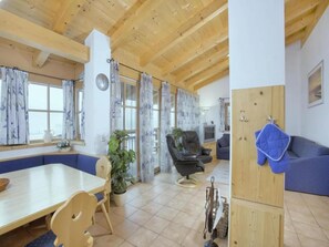 A spacious living area with a wooden ceiling, a dining table with chairs, a blue sofa, a black leather chair, and a wooden cabinet with a blue cloth.
