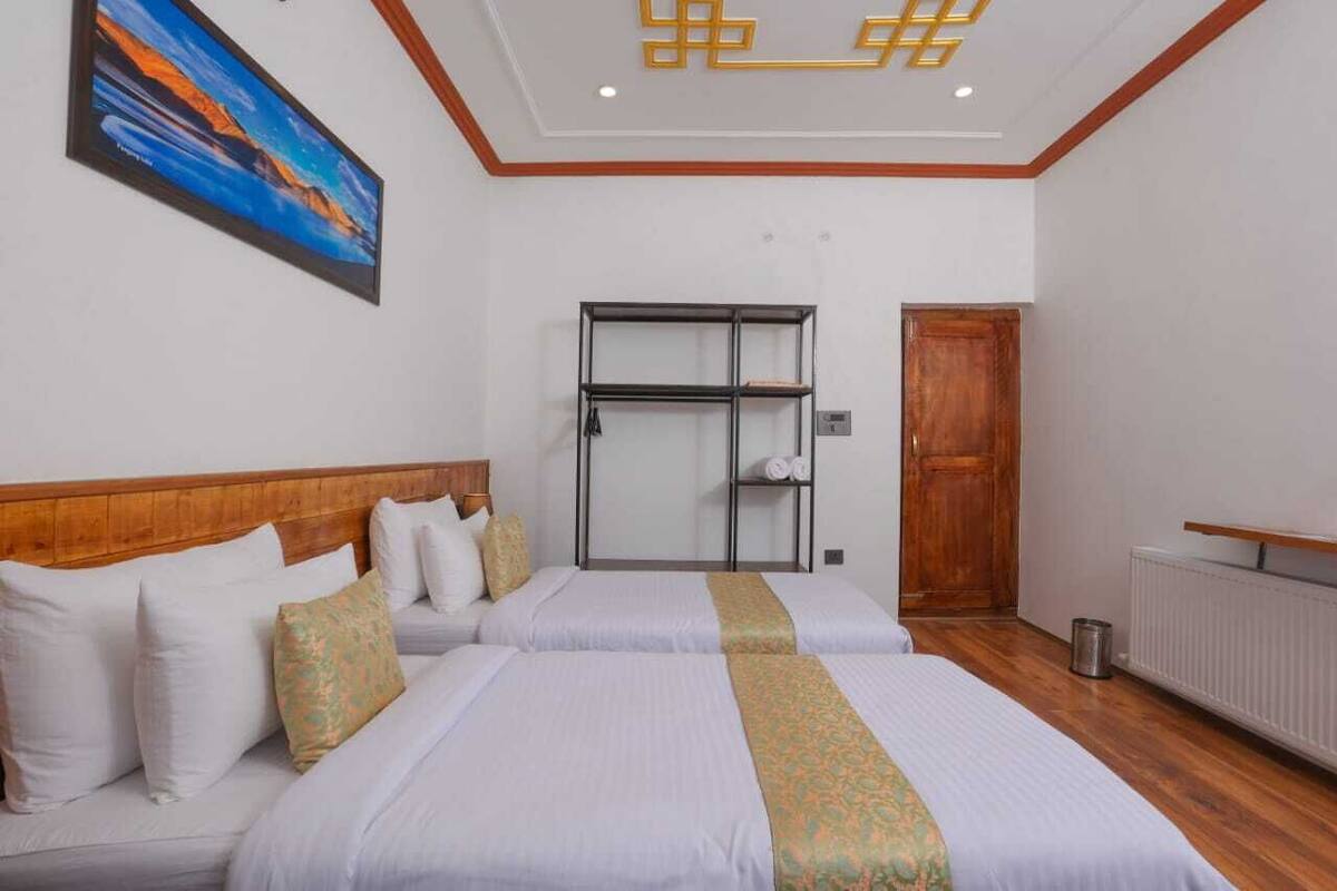 A hotel room with two beds, a wooden headboard, a painting on the wall, a metal shelf unit, and a wooden door.