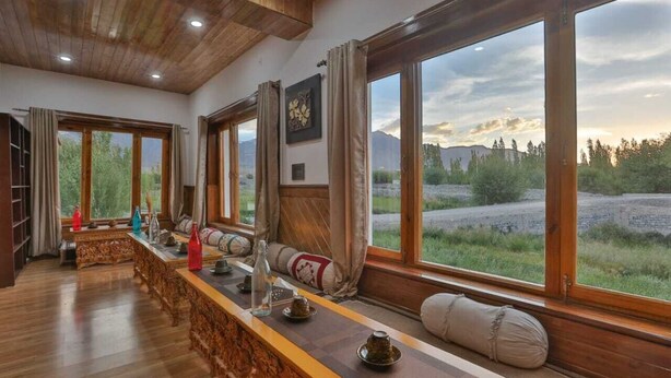 A long wooden bench with cushions and a table set for tea, large windows with curtains, and a view of a natural landscape. - Kangseng House (Leh)