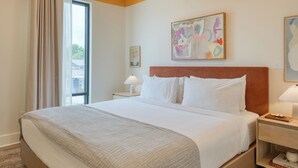 A hotel room with a large bed, a nightstand with a lamp, a small table with a book, and a window with curtains.