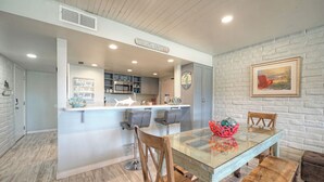 A modern kitchen with a glass dining table, wooden chairs, and a bench. There is a kitchen island with bar stools, a white brick wall, and a framed picture on the wall. - Sleeps 20 | 3 Condos w/ Beach Access, Pool, Tennis Courts | Sea Sands Complex (Port Aransas)