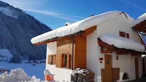 A snow-covered chalet with a wooden facade, a snowman, and a mountainous backdrop.