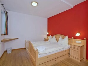 A bedroom with a wooden bed, white bedding, a red accent wall, a bedside table with a lamp, and a small wooden nightstand. - Scenic Valley Chalet in Austria (Wald-Königsleiten)