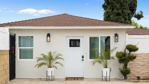 A single-story house with a white exterior, a brown tiled roof, and a front door with a small window above it. There are two potted plants on either side of the door. - Cozy & Dreamy Getaway home ! (Montebello)