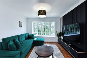 A living room with a green sofa, a round coffee table, a TV on a black stand, and a window with a view of greenery. - 2-Bed in Quiet Area, Easy City & Transport Access (Nottingham)