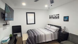 Un dormitorio con cama, ventilador de techo, televisor, mesita de noche y sofá, además de una ventana con persianas.