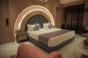 Um quarto moderno com cabeceira curvada, mesa de cabeceira, mesa lateral e uma mesa redonda com um vaso de flores.