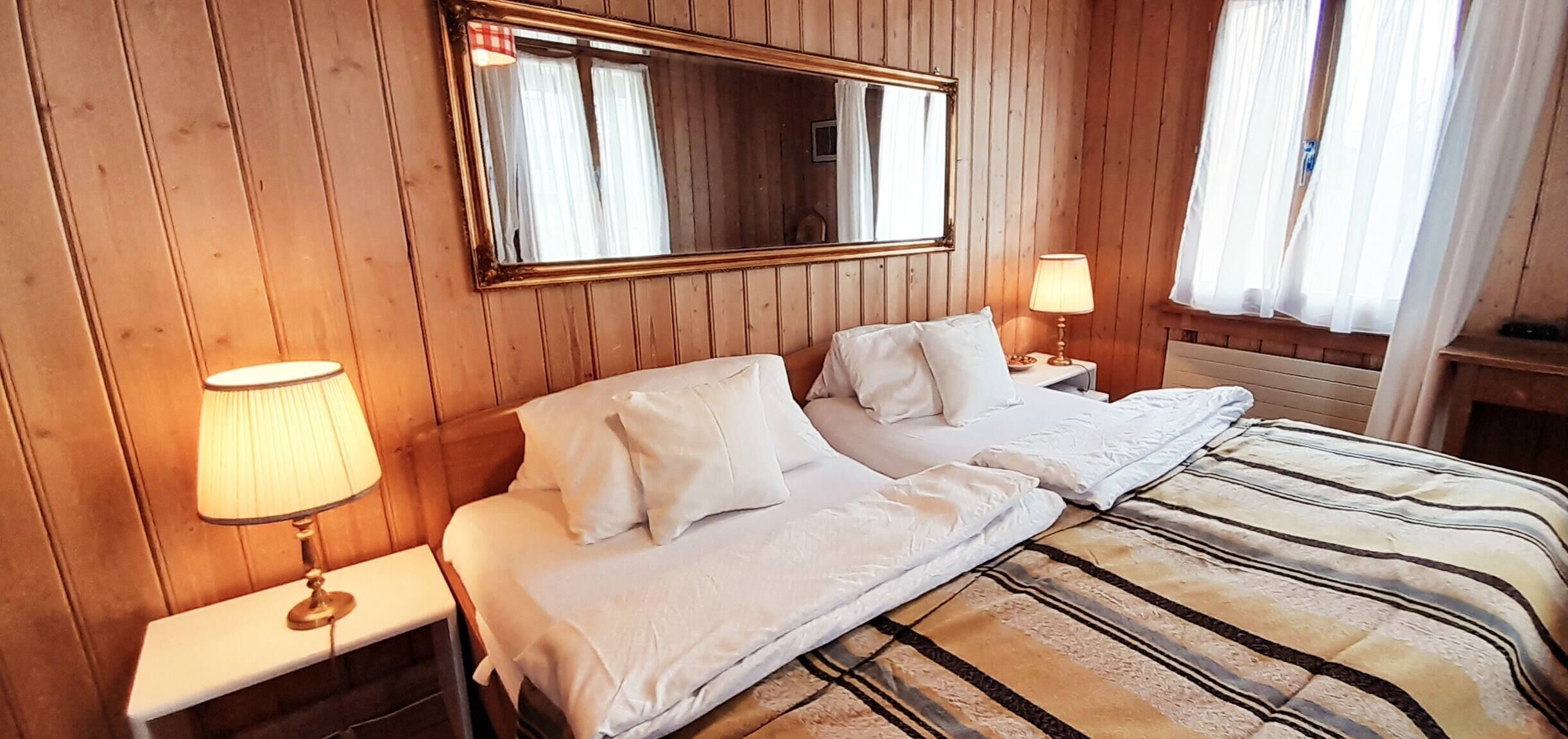A hotel room with a wooden paneled wall, a large mirror, two beds with white linens, a striped blanket, and two bedside lamps.