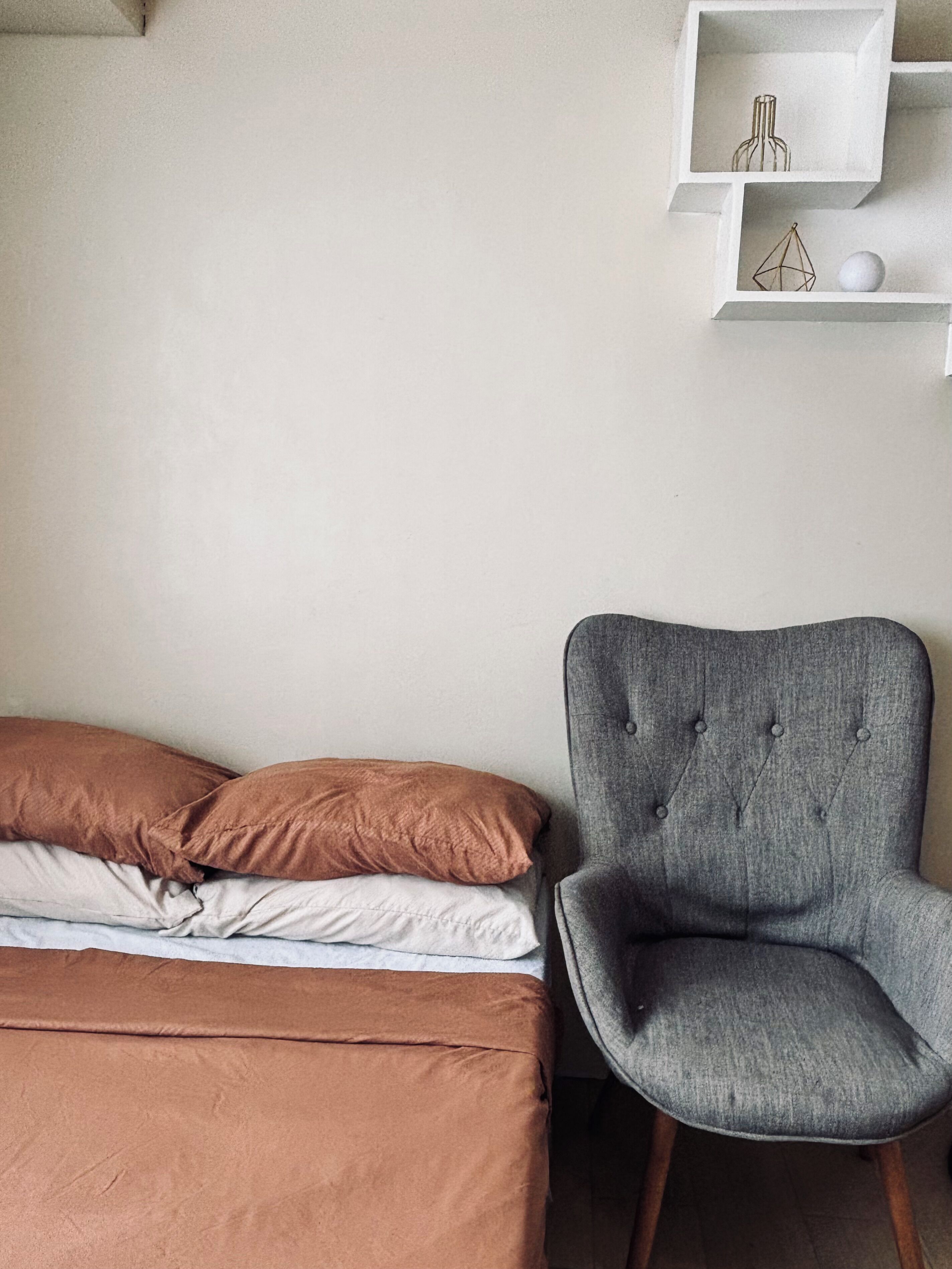 Quarto com uma cama, uma poltrona estofada cinza e uma estante branca com objetos decorativos.