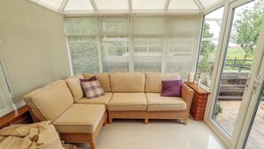 A sunroom with a sectional sofa, a wicker chair, and a view of the outdoors through large windows. - Witham View (Lincoln)