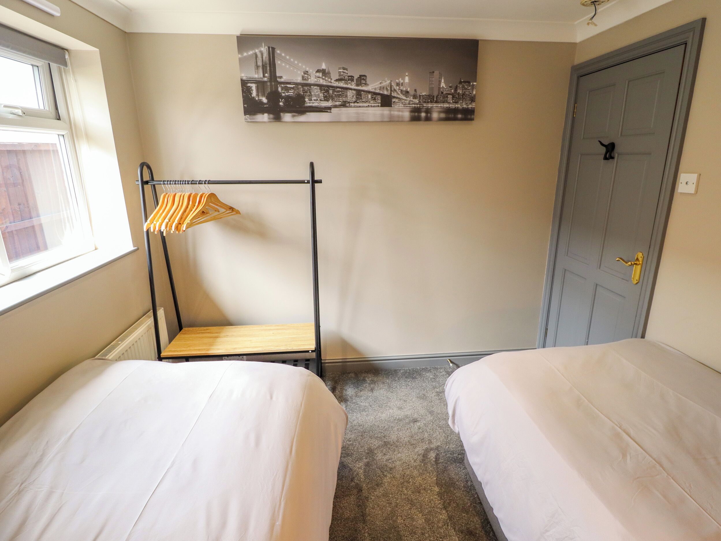 A room with two beds, a coat rack with hangers, a door, and a cityscape painting on the wall.