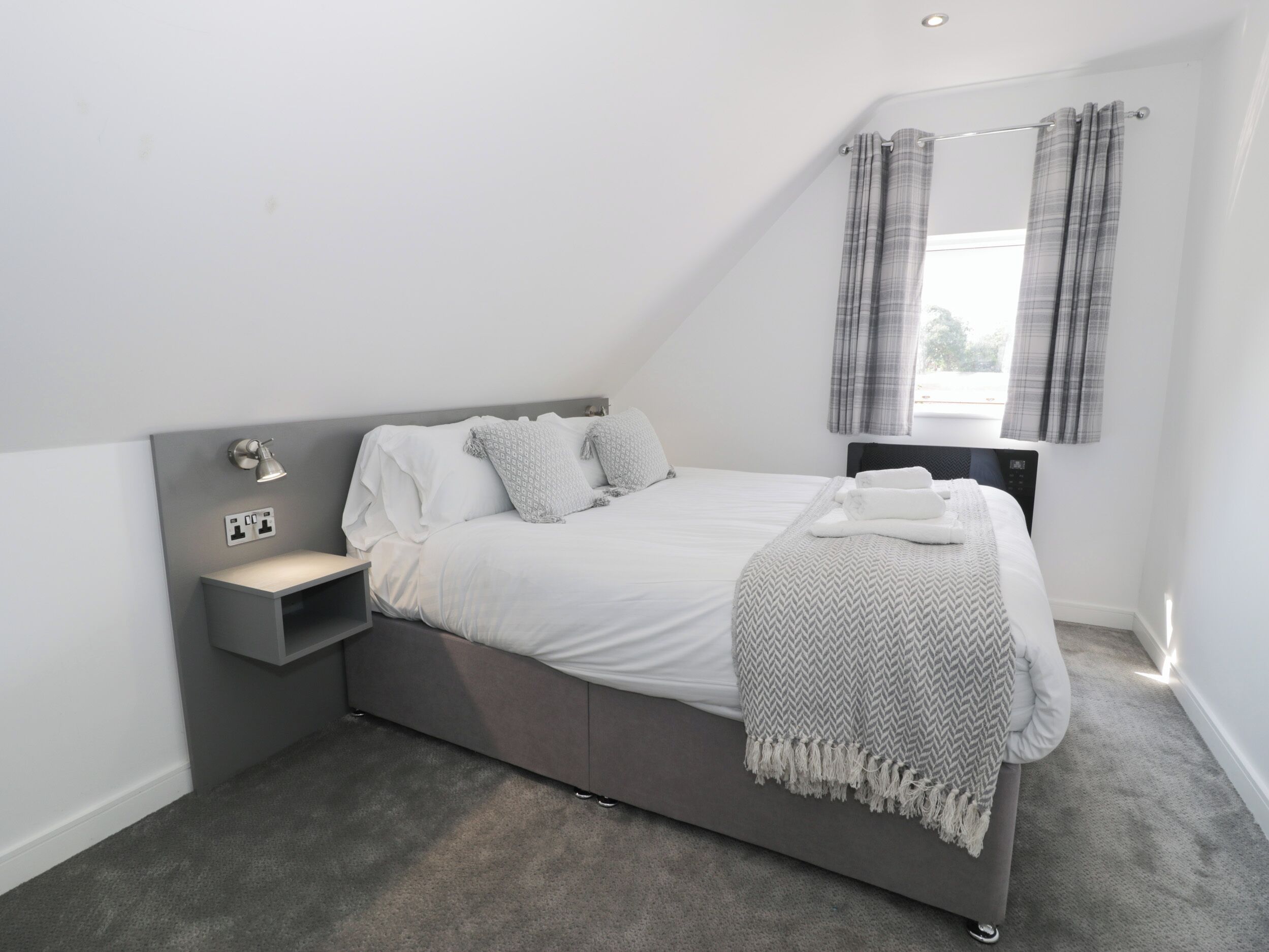 Un dormitorio moderno con una cama gris, ropa de cama blanca, una mesita de noche con lámpara y una ventana con cortinas.