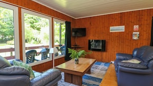 A furnished room with a wooden floor, a wooden table, two sofas, a TV mounted on the wall, and large windows with a view of greenery outside. - Lodge 1 "ash" (Isle of Arran)